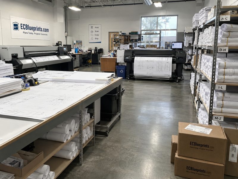 The ECBlueprints.com production floor: two large-format plotters running plan sheets, a layout table with drawings, shelves of rolled sets filed A–Z, and branded shipping boxes ready for UPS